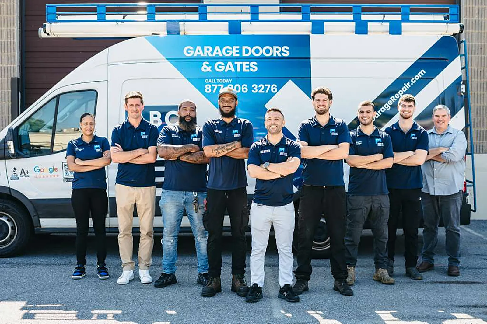 Nine garage door repair technicians in navy blue uniforms standing with arms crossed in front of a white and blue Garage Doors and Gates service van
